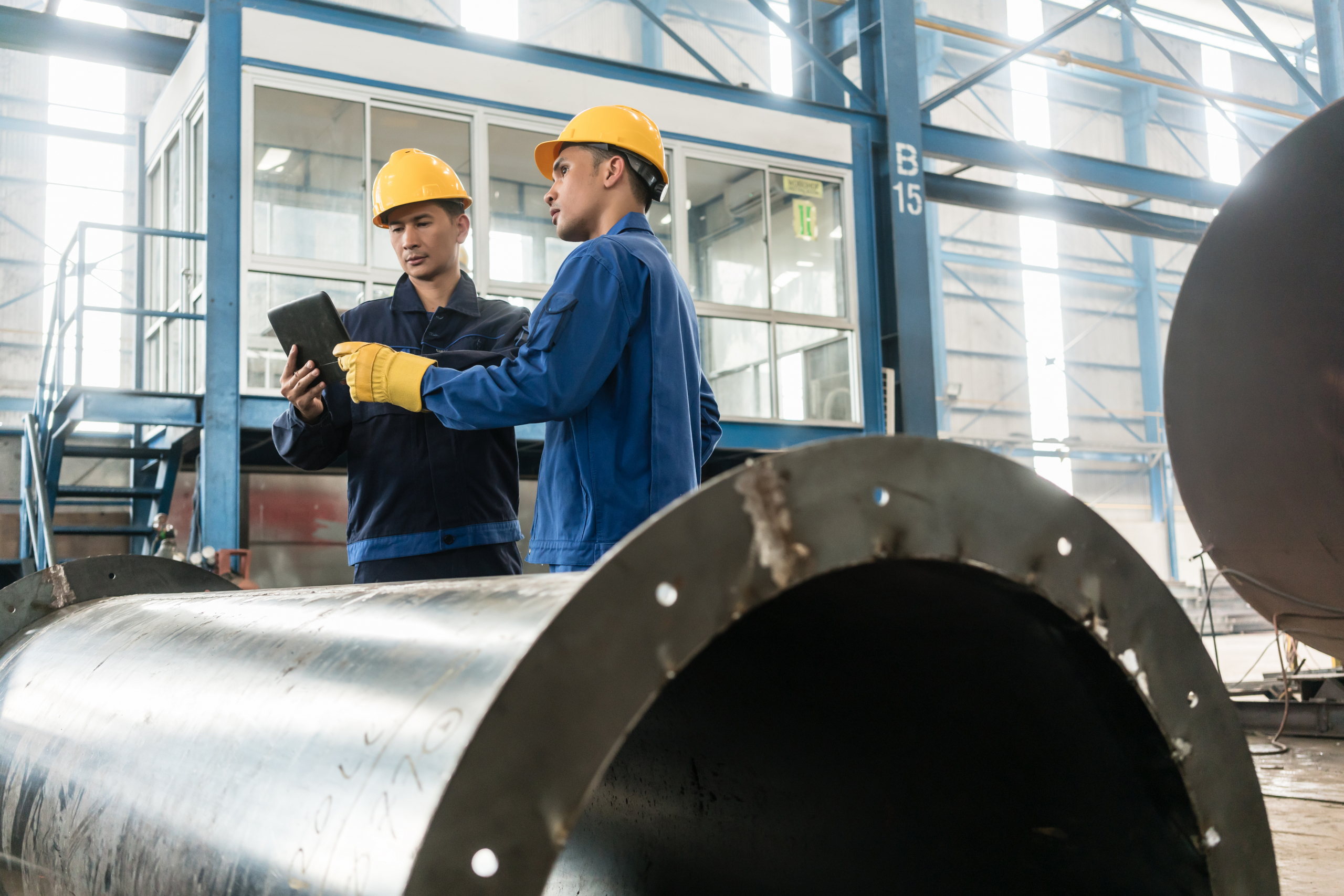 Two workers in hard hats look at a tablet in a pipe manufacturing factory